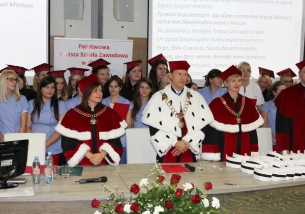 Uroczystość czepkowania licencjatów pielęgniarstwa i wręczania dyplomów, z osobami w czerwonych togach z białymi futrzanymi lamówkami na pierwszym planie i studentami w niebieskich uniformach z czerwonymi czepkami w tle.