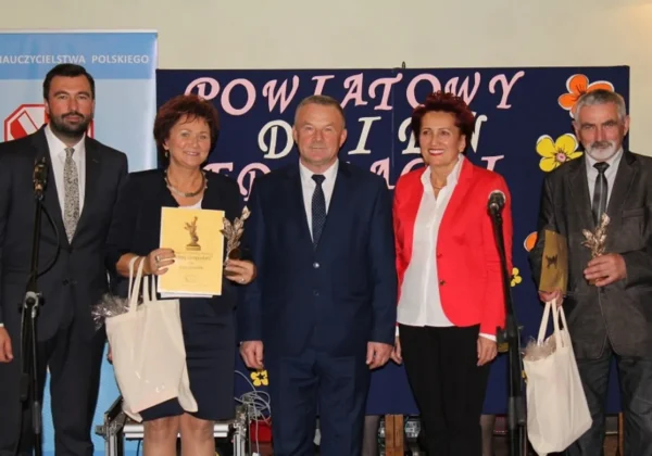 Pięć osób w formalnych ubraniach stoi na scenie podczas debaty o obronie gimnazjów, z certyfikatem i nagrodami w dłoniach, a w tle widoczny banner.