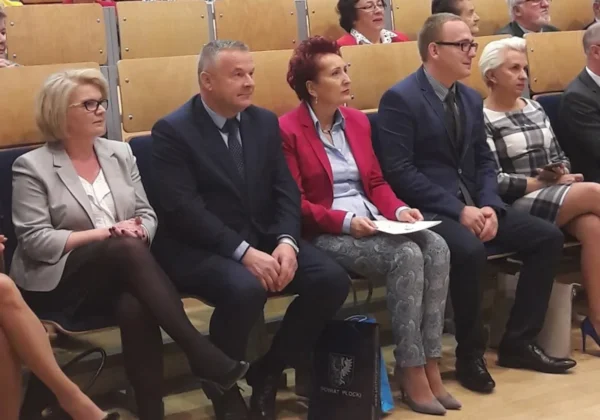 Grupa osób w formalnych ubraniach siedzi w pierwszym rzędzie sali wykładowej podczas inauguracji Roku Akademickiego Uniwersytetu Trzeciego Wieku.