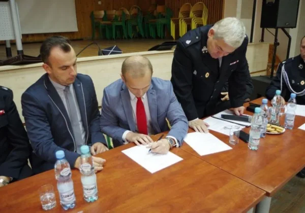 Grupa mężczyzn w garniturach i mundurach podpisuje dokumenty przy stole podczas przekazania motopomp Ochotniczym Strażom Pożarnym.