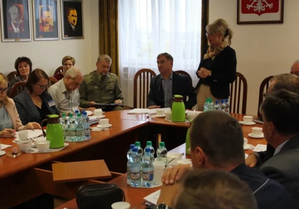 Uczestnicy ćwiczeń obronnych Dolina Wisły 2016 Powiatu Płockiego siedzą przy dużym stole w sali konferencyjnej; jedna osoba stoi i przemawia, a na stole są butelki wody, kubki i notatki.