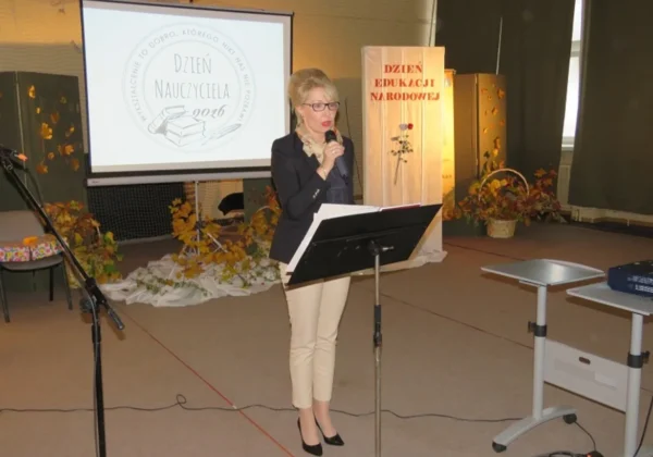 Kobieta stojąca przy podium przemawia do mikrofonu podczas Dnia Edukacji Narodowej w Zespole Szkół im. St. Staszica w Gąbinie, na tle prezentacji z logo Dzień Nauczyciela 2020 i dekoracji jesiennych.