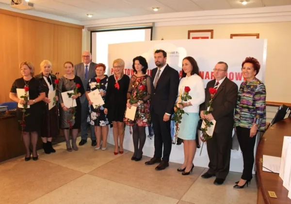 Grupa osób w eleganckich strojach stoi w rzędzie podczas XVIII Powiatowego Dnia Edukacji Narodowej, trzymając róże i dyplomy na tle baneru.