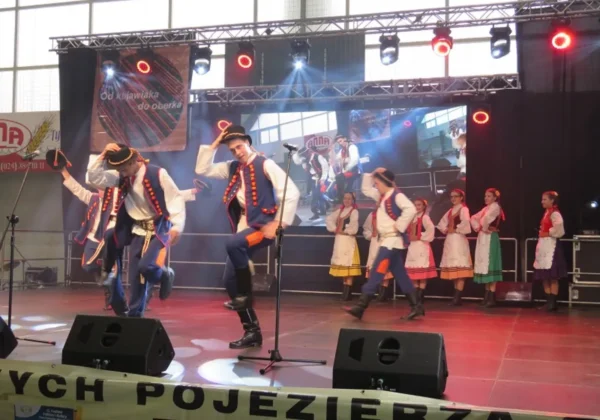 Na scenie podczas 11. Festiwalu Folkloru i Kultury Ziemi Kujawskiej i Mazowieckiej „Od Kujawiaka do Oberka” grupa tancerzy w tradycyjnych, kolorowych strojach wykonuje tańce ludowe.