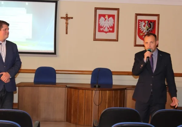 Dwóch mężczyzn w garniturach prowadzi prezentację w sali konferencyjnej; jeden przemawia do mikrofonu, drugi stoi obok, a w tle wisi projektor i dwa godła państwowe w ramkach na ścianie.