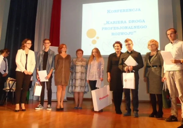 Grupa osób stoi na scenie i odbiera nagrody podczas konferencji o karierze i rozwoju zawodowym, trzymając dyplomy i torby.
