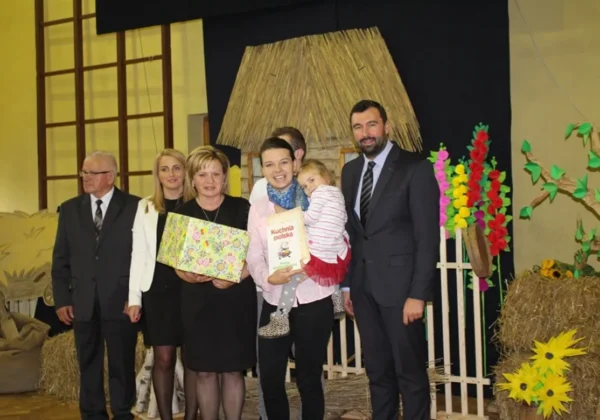 Grupa dorosłych z dzieckiem stoi na scenie podczas Dnia Chleba w ZS im. Jana Śniadeckiego w Wyszogrodzie, trzymając prezenty, w otoczeniu dekoracji z siana i kwiatów.