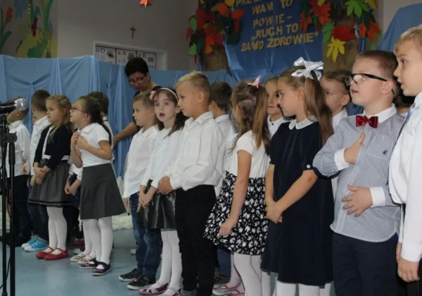 Grupa przedszkolaków ubranych na galowo stoi w szeregu na scenie podczas szkolnego występu, z bannerem „Ruch to zdrowie” w tle.