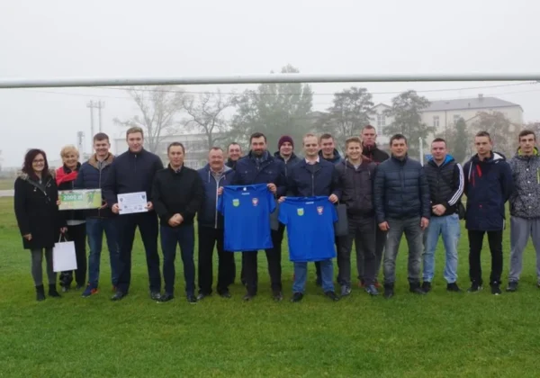 Grupa osób na boisku sportowym w Radzanowie stoi przed bramką, prezentując niebieskie koszulki i dokumenty związane z modernizacją obiektu.