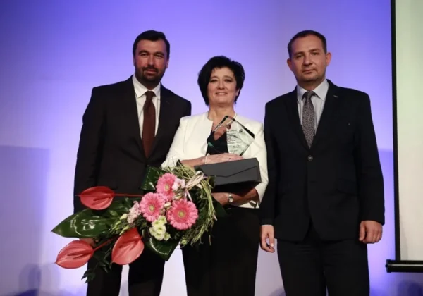 Trzy osoby stoją na scenie podczas jubileuszu 25-lecia Okręgowej Izby Pielęgniarek i Położnych Regionu Płockiego; kobieta w białej marynarce trzyma nagrodę i bukiet kwiatów, flankowana przez dwóch mężczyzn w garniturach.