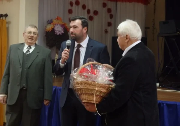 Trzech mężczyzn na scenie podczas Dnia Seniora w Bielsku; jeden mówi do mikrofonu, drugi stoi obok, a trzeci trzyma kosz z prezentem owiniętym folią.