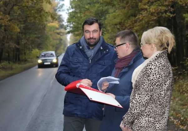 Trzy osoby stoją na jesiennym lesie przy mokrej drodze podczas inspekcji modernizacji drogi Soczewka – Łąck; jedna trzyma czerwoną teczkę z dokumentami, a dwie omawiają materiały, podczas gdy w tle nadjeżdża samochód.