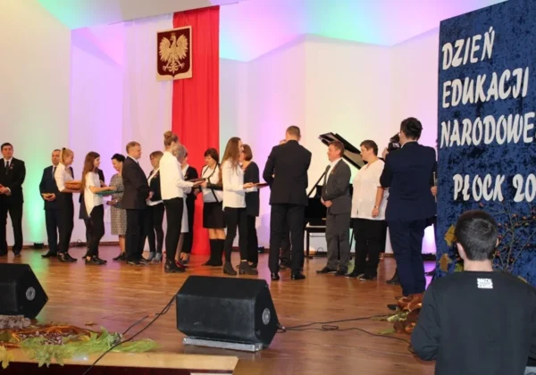 Grupa osób na scenie wręcza nagrody podczas obchodów Dnia Edukacji Narodowej w Płocku, z polską flagą i napisem Dzień Edukacji Narodowej Płock 2016 w tle.