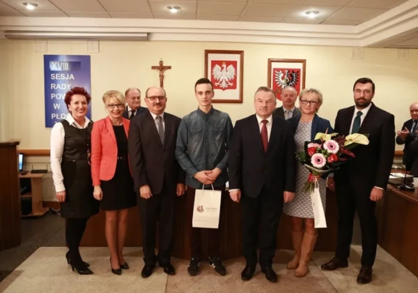 Grupa osób w formalnych strojach stoi w sali obrad podczas XVIII Sesji Rady Powiatu w Powiecie Płockim; dwie osoby trzymają bukiety kwiatów, a w tle widać krzyż i dwa polskie herby.