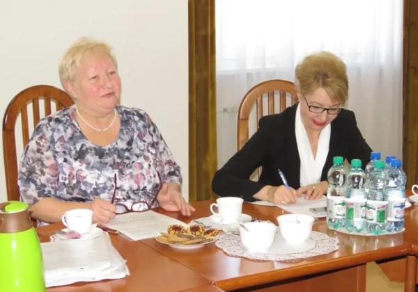 Dwie kobiety siedzą przy stole podczas spotkania o pomocy żywnościowej dla potrzebujących mieszkańców; jedna zapisuje na dokumentach, druga uśmiecha się, a na stole są napoje, filiżanki i ciasteczka.