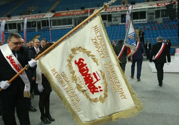 Starosta i Przewodniczący Rady Powiatu niosą sztandar Solidarności podczas zjazdu, na tle hali z widownią i innymi uczestnikami w garniturach.