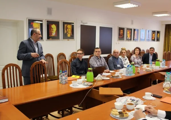 Mężczyzna w garniturze prowadzi szkolenie formacyjne OC podczas spotkania w sali konferencyjnej, a uczestnicy siedzą przy długim stole z notatkami, napojami i przekąskami.