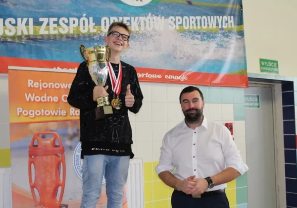 Młody pływak stojący na podium z dużym pucharem i medalami, uśmiechnięty, podczas ceremonii wręczania nagród obok niego dorosły mężczyzna w białej koszuli.