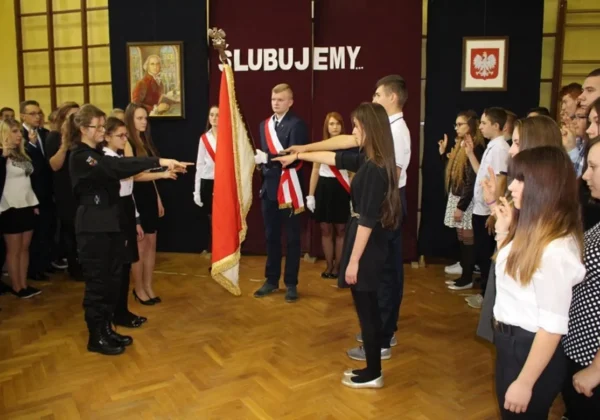 Uczniowie w galowych strojach stoją w dwóch rzędach podczas ceremonii ślubowania przy sztandarze na wysogrodzkich uroczystościach.