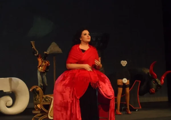 Kobieta ubrana w efektowną, czerwoną suknię siedzi na scenie teatralnej w otoczeniu rekwizytów i dekoracji.