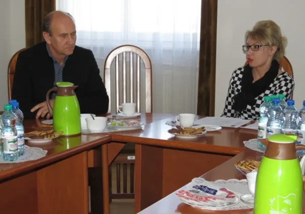 Dwóch dorosłych uczestników spotkania siedzi przy stole w sali konferencyjnej, rozmawiając; na stole znajdują się zielone dzbanki termiczne, butelki wody, filiżanki i talerze z ciasteczkami.
