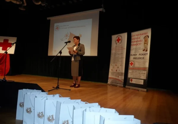 Kobieta w garniturze przemawia do mikrofonu na scenie podczas prezentacji; w tle projekt na ekranie, po prawej stoją banery Polskiego Czerwonego Krzyża, a na podłodze w pierwszym planie leżą jasnoniebieskie torby z godłem.