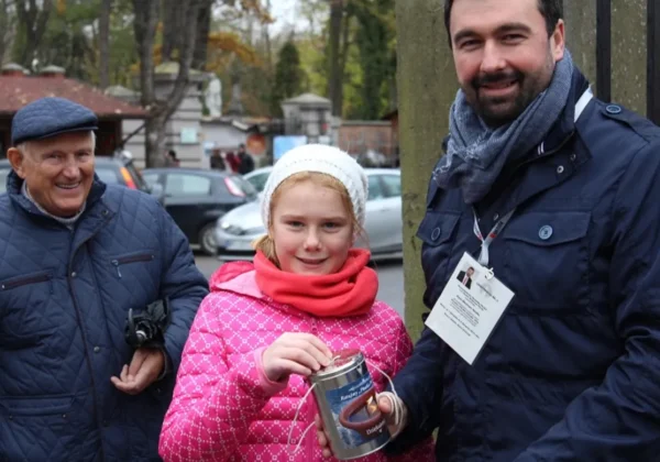 Trójka ludzi na ulicy podczas akcji charytatywnej „Ratujmy Płockie Powązki”: starszy mężczyzna w granatowej kurtce, dziewczynka w różowej kurtce z białą czapką wrzuca pieniądze do puszki, obok stoi młody mężczyzna z identyfikatorem.