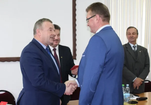 Dwóch mężczyzn w garniturach ściska sobie ręce podczas ceremonii odznaczeń Zasłużeni dla Powiatu Płockiego w Nowym Duninowie, w tle stoją inni uczestnicy.