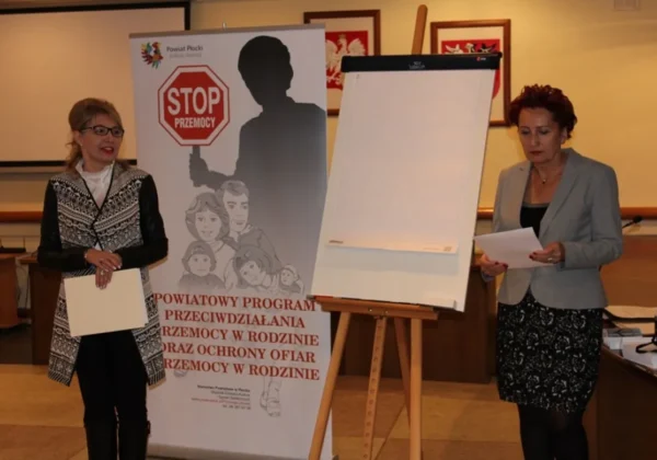 Dwie kobiety stoją przy banerze STOP PRZEMOCY i tablicy do prezentacji, prezentując materiały dotyczące przeciwdziałania przemocy w rodzinie.