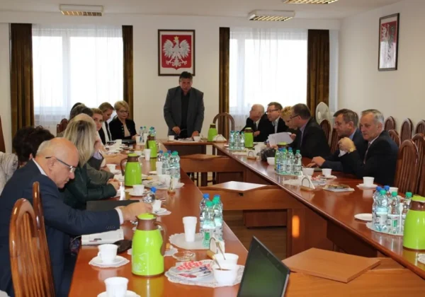 Uczestnicy spotkania w sali konferencyjnej omawiają afrykański pomór świń i problem grzebowisk, a prowadzący stoi przy stole, z tyłu widoczny jest polski herb.