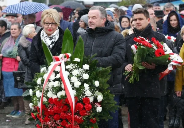 Na pierwszym planie para trzyma duży czerwono-biały wieniec z kwiatów podczas obchodów Narodowego Święta Niepodległości 11 listopada, wśród tłumu w zimowej pogodzie.