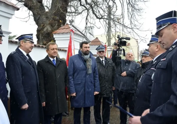 Grupa mężczyzn w mundurach i eleganckich płaszczach stoi w zimowej scenerii przed flagami podczas obchodów Narodowego Święta Niepodległości w Wyszogrodzie; pada śnieg.
