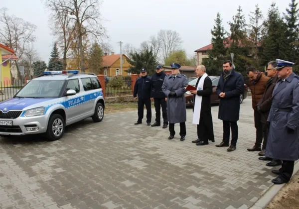 Nowy radiowóz policji stoi na placu wyłożonym kostką brukową w Bodzanowie, otoczony policjantami i duchownym podczas uroczystości.