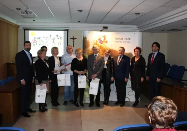 Grupa osób w formalnych ubraniach stoi na sali konferencyjnej podczas podsumowania współzawodnictwa sportowego szkół, trzymając białe torby z logo powiatu.