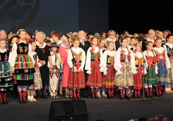 Grupa dzieci w kolorowych strojach ludowych stoi na scenie podczas jubileuszu 70-lecia działalności Dzieci Płocka.