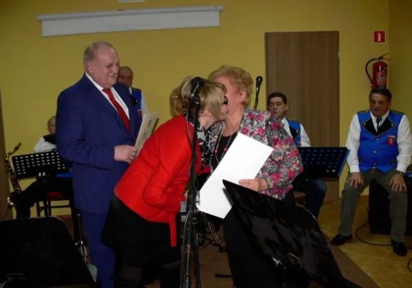 Uroczystość z okazji 5-lecia Koła Gospodyń Wiejskich w Podlasiu: dwie kobiety składają sobie pocałunek na powitanie przy mikrofonie, podczas gdy mężczyzna w garniturze wręcza dokument, a w tle grają muzycy.