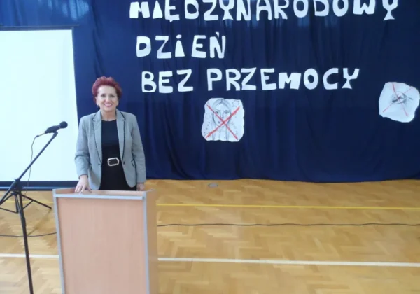 Kobieta stoi przy mównicy na sali gimnastycznej, a za nią duży baner z napisem „Międzynarodowy Dzień Bez Przemocy”.