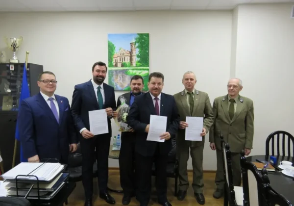 Grupa sześciu mężczyzn w garniturach i mundurach wojskowych stoi w biurze, prezentując podpisane dokumenty.