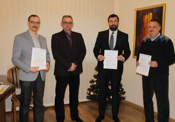 Czterech mężczyzn w garniturach stoi w biurze, każdy trzyma kartkę z dokumentem, a w tle wisi obraz i stoi choinka.