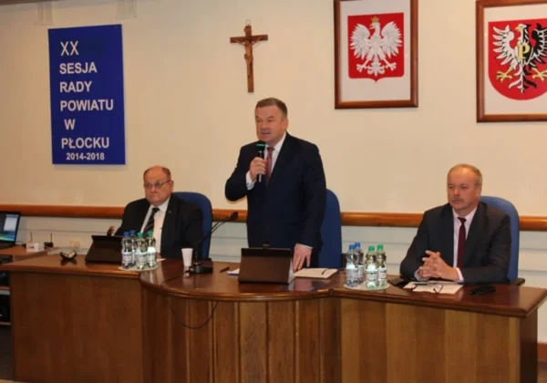 Trzech mężczyzn w garniturach przy dużym drewnianym stole podczas sesji Rady Powiatu Płockiego; jeden stoi i przemawia do mikrofonu, a w tle widoczne są krzyż, herby i baner XX Sesja Rady Powiatu w Płocku.