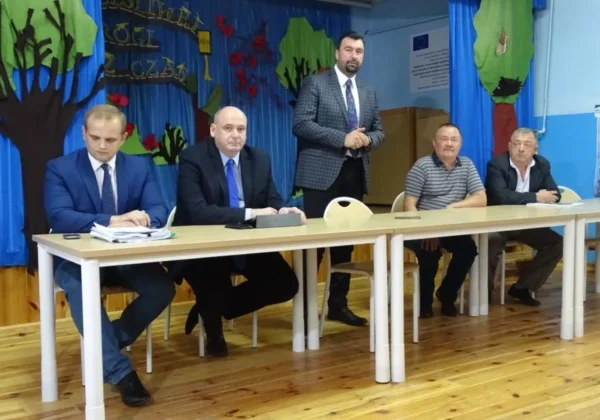 Pięciu mężczyzn w garniturach siedzi przy długim stole podczas formalnego spotkania w sali z niebieskimi zasłonami i drzewiastymi dekoracjami; jeden z nich stoi.