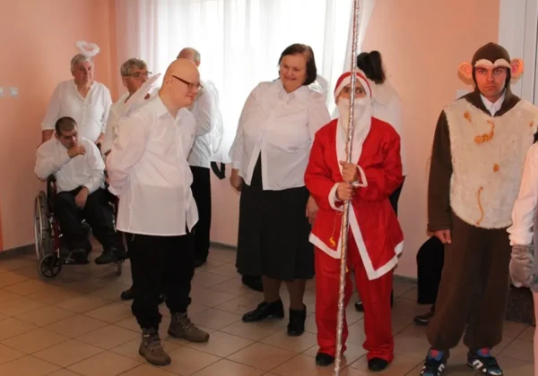 Grupa osób w białych koszulach stoi w korytarzu podczas wigilii w Goślicach, wśród nich osoba przebrana za Mikołaja z różdżką oraz osoba w stroju małpy.
