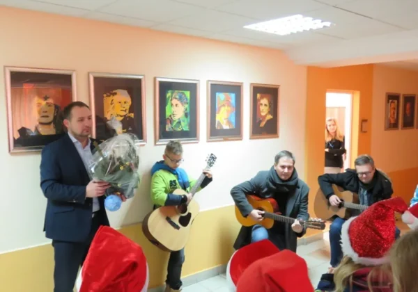 Kolędnicy i muzycy grający kolędy w holu Starostwa Powiatowego, otoczeni widownią w czerwonych czapkach świątecznych.