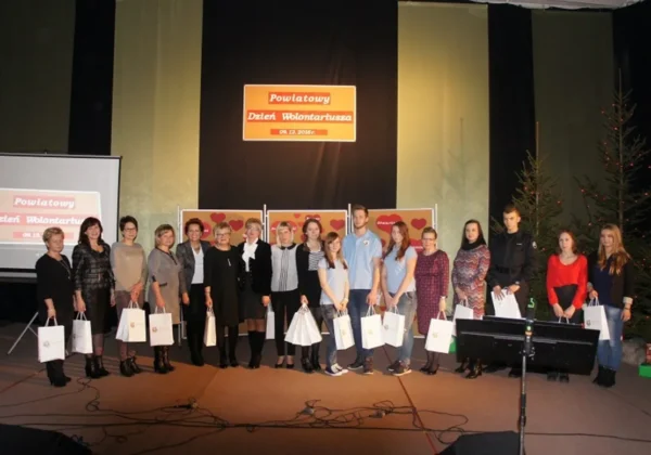 Grupa osób stoi na scenie podczas uroczystości Powiatowy Dzień Wolontariusza, wręczając prezenty w białych torbach, z widocznym plakatem i świątecznymi choinkami w tle.