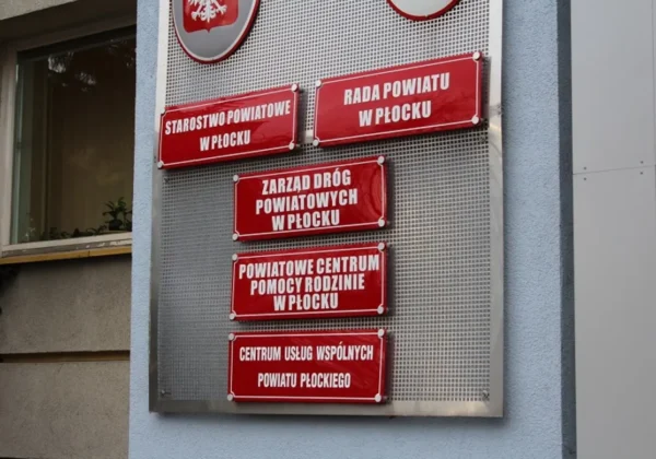 Na metalowej tablicy przy budynku widać pięć czerwonych tabliczek z nazwami instytucji powiatowych w Płocku: Starostwo Powiatowe w Płocku, Rada Powiatu w Płocku, Zarząd Dróg Powiatowych w Płocku, Powiatowe Centrum Pomocy Rodzinie w Płocku i Centrum Usług Wspólnych Powiatu Płockiego.