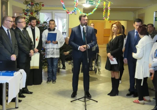 Mężczyzna w garniturze przemawia do zgromadzonych podczas wigilijnego spotkania w Brwilnie, obok stoi duchowny i inni goście przy udekorowanej choince i kolorowych ozdobach.