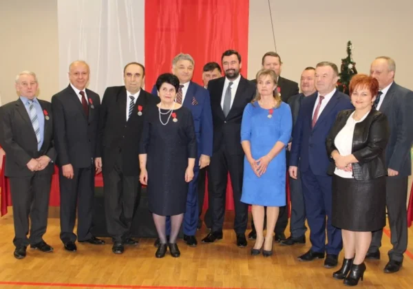 Grupa osób w garniturach i eleganckich sukniach, część z medalami na piersiach, stoi na scenie podczas uroczystości Zasłużeni dla Powiatu Płockiego w Gminie Staroźreby, z flagą Polski w tle.