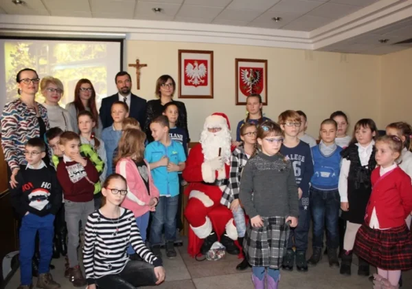 Grupa dzieci i dorosłych stoi w sali Starostwa wokół siedzącego Świętego Mikołaja; w tle widoczne krzyż i dwa herby.