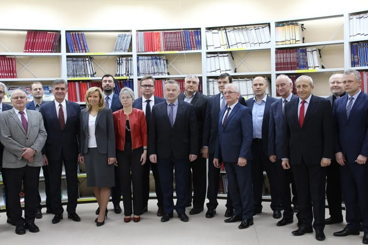 Grupa urzędników i radnych w garniturach stoi razem w biblioteczce technicznej przed regałami z teczkami; wśród nich znajdują się starosta i przewodniczący Rady przy Politechnice.