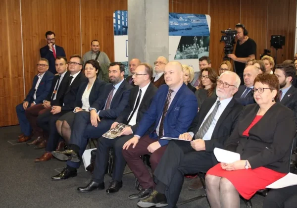 Grupa osób w garniturach siedzi w sali konferencyjnej podczas spotkania o funduszach unijnych, a operator kamery nagrywa wydarzenie.